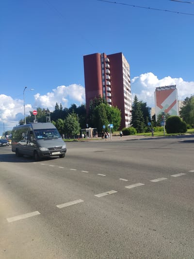 A gray Mercedes-Benz Sprinter van with the license plate AM5568-2 is driving on a paved road in Navapolatsk, Belarus. The van is in the foreground, partially obscuring a pedestrian crossing. In the background, there are two tall apartment buildings, one brick-red and the other with a colorful geometric pattern. Trees line the street, and fluffy white clouds are scattered across a clear blue sky. Several pedestrians can be seen on the sidewalk near the buildings. A traffic light is visible, and there are street signs and a traffic pole. The scene appears to be an intersection on a sunny day.