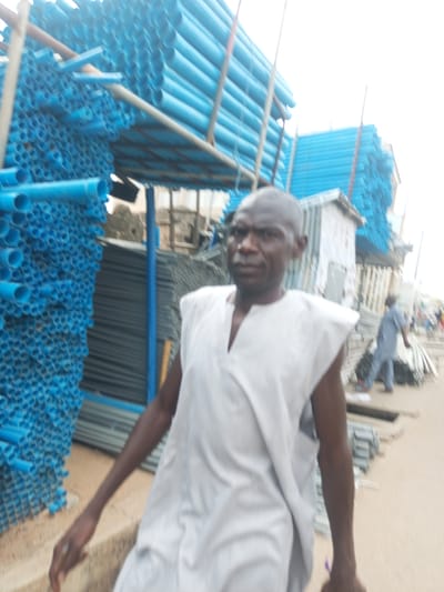 In Arawa, Nigeria, a man in a white garment stands in a market or storage area filled with blue pipes. He is the primary subject, positioned in the foreground, looking towards the viewer. Behind him, the scene is blurred but suggests an outdoor environment with stalls or structures. Large stacks of bright blue pipes are a dominant feature, arranged in rows and forming a significant part of the visual landscape. To the right and further in the background, other individuals are visible, also appearing to be in a commercial or industrial setting. The ground is covered with a light-colored surface, possibly sand or dirt, and there are more stacks of what appear to be metal or PVC materials. The overall atmosphere suggests a bustling, outdoor marketplace where construction materials are sold or stored.