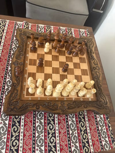 The image displays an indoor scene centered on a wooden chess set. The specific location in Aragats, Armenia, cannot be determined from the visual evidence.

The primary subject is a complete chess set, featuring a carved wooden board and chess pieces. The board's playing surface consists of inlaid light and dark wood squares, with algebraic notation (A-H, 1-8) visible on its edges. A wide, dark brown wooden frame, intricately relief-carved with decorative floral or scroll motifs, surrounds the playing area.

The chess pieces are made of wood; white pieces are light-colored, and black pieces are dark-colored. The pieces are arranged as if in an ongoing game.
- White's pieces, positioned at the bottom of the board, include rooks on a1 and h1, knights on b1 and g1, bishops on c1 and f1, a queen on d1, and a king on e1. White pawns are located on a2, b2, c2, d2, f2, g2, h2, and one pawn is advanced to e4.
- Black's pieces, positioned at the top of the board, include rooks on a8 and h8, knights on b8 and g8, bishops on c8 and f8, a queen on d8, and a king on e8. Black pawns are located on a7, b7, c7, e7, f7, h7, and two pawns are advanced to d5 and g6.

The chess board rests on a patterned fabric, likely a table runner, which exhibits vertical stripes with repeating geometric and curvilinear designs in red, black, off-white, and dark blue/grey. This fabric covers a section of a dark brown wooden table surface. In the upper background, a portion of a grey upholstered piece of furniture, possibly an armrest or backrest of a sofa or chair, with dark trim, is visible. The scene is static, depicting no individuals or active interactions.