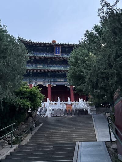 The image depicts a traditional Chinese building partially obscured by trees, viewed from a low angle looking up. The building has a multi-tiered roof with blue and black decorative elements, red columns, and a blue sign with gold lettering in Chinese characters. A person is visible on the stairs near the building, which has white balustrades. The stairs are concrete and lead upwards, alongside a metal railing. The environment appears to be a park or garden setting in daylight. Based on the style of architecture, the building is likely located in the Xicheng District, China.