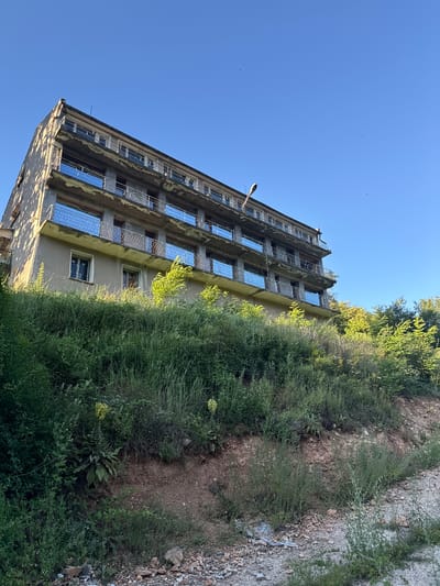 An abandoned, multi-story building with a weathered concrete facade stands on a hillside covered in dense green vegetation. The building has multiple balconies with reflective blue panels. The sky above is clear and blue. The foreground shows a rough, gravelly path leading up to the overgrown hillside. The scene appears to be located in Shipkovo, Bulgaria.