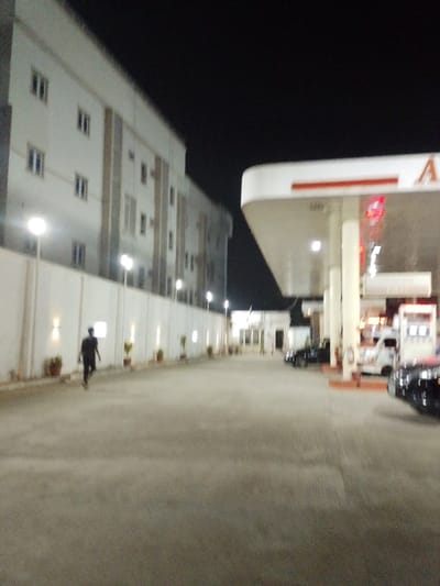 This image depicts a nighttime scene at a fueling station and adjacent structures in Babban Giji, Nigeria. The foreground features a wide, paved forecourt. To the right, a white gas station canopy with a red border extends over multiple fuel pumps, which display red digital readouts. Several vehicles, including dark-colored cars and a white van, are parked beneath the canopy. To the left, a long, illuminated white wall runs parallel to the paved area, featuring regularly spaced wall-mounted lights and potted plants at its base. Behind this wall, a multi-story building with a light-colored facade and multiple rectangular windows is visible. A single individual, dressed in dark clothing, is observed walking from left to right on the paved surface alongside the white wall. The entire scene is brightly illuminated by artificial lights against a dark night sky.