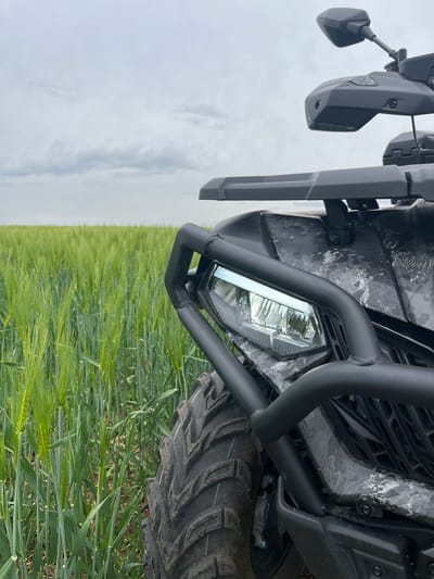 The image depicts the front of an off-road vehicle parked in a field of green grain crops, likely wheat or barley. The vehicle is oriented diagonally, with the front headlight and a black, tubular bumper guard prominently visible. The tire is visible to the right, partially obscured by the crops. A rearview mirror is visible on the left side of the image. The vehicle has a camouflage pattern on its body. The background consists of a vast field of green crops stretching to the horizon, topped by an overcast sky. Given the presence of grain crops, the location is likely rural. While precise location in Golyanovtsi, Bulgaria is impossible to determine from the image, it suggests a rural setting with agricultural fields.