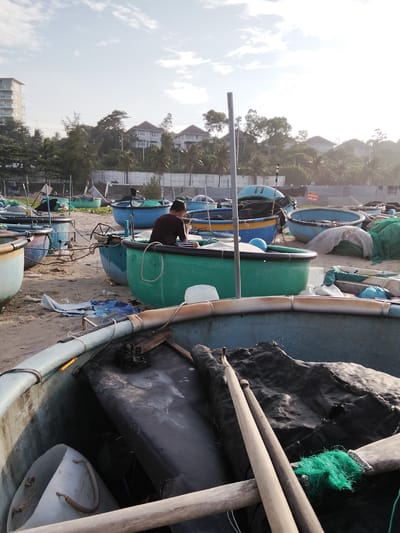 In Phú Thủy, Vietnam, a person sits in a round, turquoise fishing boat on a sandy beach. The person, wearing a maroon shirt, is looking down at their hands. The boat contains a black tarp and some wooden oars. Many similar boats are present on the beach, some containing nets or other equipment. In the background, there are palm trees, houses, and a modern building. The sky is partly cloudy.