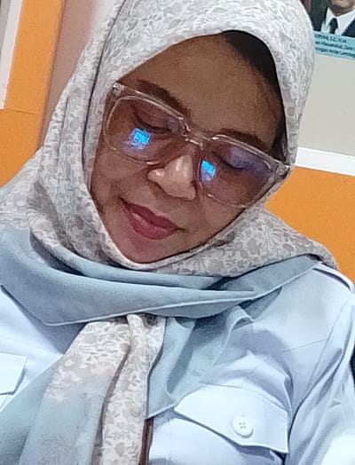The image shows a close-up, waist-up shot of a woman wearing a light blue uniform shirt, a light blue hijab, and a patterned headscarf. She is also wearing large, clear-framed glasses. Her eyes are closed, and she has on dark lipstick. The background appears to be an orange wall. Based on the context provided, the location is Meunasahaluedrienlhoksukon Barat, Indonesia. There is a faint image of a man in a suit and tie visible on the wall behind the woman.