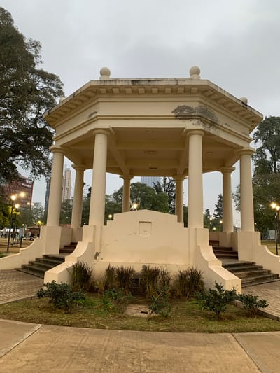 This image depicts an octagonal, cream-colored gazebo situated in a public park in Cordoba, Argentina, under an overcast sky. The gazebo features a flat roof with a decorative frieze and four spherical finials on its corners. It is supported by eight cylindrical columns. Two sets of dark-colored stairs lead up to the structure's platform from opposite sides, integrated into a curved, cream-colored base. The structure exhibits signs of wear, including peeling paint, discoloration, and dark stains on its upper facade and roofline.
The immediate surroundings of the gazebo include patches of green grass, several small green shrubs, and taller, dry ornamental grasses planted around its base. Paved pathways of interlocking bricks lead to the gazebo, with a lighter concrete path visible in the immediate foreground.
In the background, mature trees with dense foliage frame the gazebo on both sides. Beyond the trees, an urban landscape is visible, featuring several tall buildings. A prominent, light-colored skyscraper stands in the central background, and to the left, a building with a reddish-orange facade displaying the "Naranja X" logo is discernible. Multiple streetlights with glowing warm yellow/orange lights are active throughout the park, indicating low natural light conditions, possibly dusk, dawn, or an heavily overcast day. A few indistinct human figures are visible on a paved area in the distant left background.
