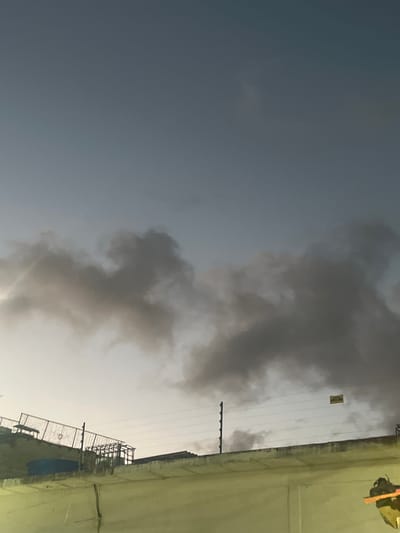 The image shows a partially cloudy sky with dark grey clouds dominating the upper portion. Below the clouds, there is a wall and structures that appear to be part of a construction site or industrial area. A fence with visible wires runs along the top of the wall, and a sign with yellow text is attached to the wires. The lighting suggests it is either early morning or late evening. No people are visible in the image. The overall scene is urban or industrial, with a sense of unfinished construction or an active work area. The location of Conuco Viejo, Venezuela is not discernible from the visual elements in the image itself.