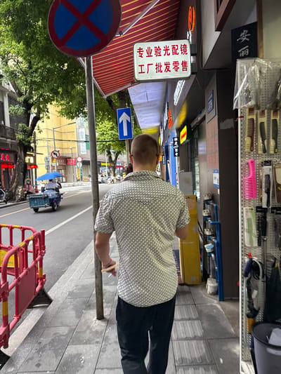 A man with short, light brown hair walks away from the camera on a sidewalk in Liwan District, China. He is wearing a short-sleeved white shirt with a black geometric pattern and black pants. He holds a cigarette in his right hand. The sidewalk is made of concrete tiles and has some debris on it. A red and yellow barricade is on the left side of the sidewalk. A street with a blue tricycle is visible to the left of the sidewalk. A sign with Chinese characters is mounted on the side of a building on the right, with rows of combs and hairbrushes displayed on a rack in front of the building. A blue and red "no parking" sign is visible in the background.