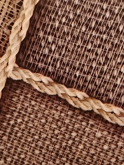 This close-up image, located in the city of Biliaivka, Ukraine, presents a detailed view of two distinct textile elements. A thick, light-beige braided rope, exhibiting a visibly fuzzy texture consistent with natural fibers, runs diagonally across the frame in a gentle 'S' curve. The rope partially overlays a darker, patterned textile background. This background fabric features a grid of fine, light-brown lines, within which numerous small, irregularly shaped, lighter elements resembling leaves or petals are densely arranged, creating a textured, decorative pattern. The scene is devoid of any human presence, subjects, actions, or interactions, focusing exclusively on the tactile qualities and interplay of the textile materials under even, diffused lighting conditions.