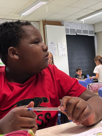 This image, captured in a classroom setting in La Fouillouse, France, features a person in the foreground, seen from a low, tilted angle. The main subject has dark skin and short, curly dark hair, wearing a red t-shirt adorned with a black, stylized animal logo on the chest, possibly a meerkat. They are holding a clear ballpoint pen in their right hand, with a focused gaze directed upwards and to the left. A small silver stud earring is visible in their left ear.
In the mid-ground, other individuals are present: a person with fair skin and glasses is partially visible to the right, and further back, a young girl in a white t-shirt stands near a blue chair, beside a pink, sparkly bag. Behind her, a boy in a black t-shirt is seated, looking towards the camera with a slight smile.
The setting is an educational environment with light-colored walls, white acoustic panels or radiators, and fluorescent lighting. A large black chalkboard is prominent in the background. Numerous papers are taped to the walls, some displaying text. Notably, one chart is titled "Présent - Prétérit" (Present - Preterite), indicating a French language lesson. Another paper asks "WHAT'S YOUR VALUE?". The main subject is at a light-colored desk, where a colorful paper is also visible. The scene suggests a classroom during a learning or study session, with students engaged in their work.