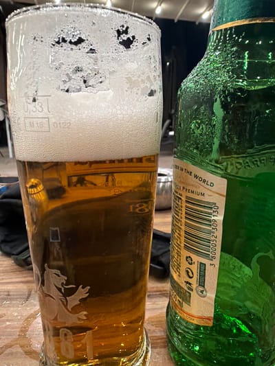 The image presents a still life of a green beer bottle and a glass of beer, likely in an indoor setting such as a bar or restaurant in Markovo, Bulgaria. The background is dark and somewhat blurred, with soft lights visible in the upper left, suggesting an evening ambiance.

The green glass bottle, positioned above the beer glass, is covered in condensation, indicating it is cold. A paper label wraps around the bottle, displaying a barcode "3 800052 309 130". Visible text on the label includes phrases such as "BEER IN THE WORLD" and "PREMIUM", the volume "500 ml", and the website "MEN.COM". Below these, a consumer helpline number "0700 13 880" is printed, preceded by "ТЕЛЕФОН НА ПОТРЕБИТЕЛЯ" (Consumer's phone in Bulgarian). The bottle's glass features an embossed, stylized animal figure, possibly a lion.

Below the bottle, a clear glass holds a golden-amber beer, topped with a substantial head of white, foamy beer. The glass also shows condensation on its exterior. An etched design of a stylized deer or moose is present on the side of the glass. Near the foam, the volume "0.33l" and a code "M18-0122" are visible. The items rest on what appears to be a wooden table surface.