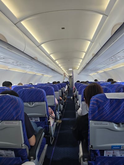 The image shows the interior of an airplane cabin, likely in or around Xiaoshan District, China, given the prevalence of Chinese text on the aircraft. Rows of blue seats with a striped pattern are visible, occupied by passengers. The aisle is carpeted in dark blue. Overhead compartments are closed, and lighting strips illuminate the ceiling. The perspective is from a seated position, looking down the aisle towards the front of the aircraft. Some passengers are looking forward, while others are facing away. The overall environment appears to be a typical commercial flight setting.