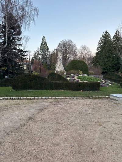 A park-like setting with a stone path along the right. Well-manicured hedges and shrubs line the path. A statue stands among bare trees in the center-left. Evergreen trees are visible in the background. The sky is light blue.