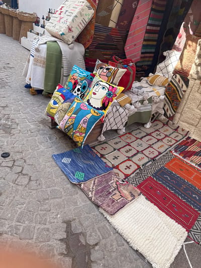 The image depicts a vibrant market stall in Essaouira, Morocco, showcasing a colorful array of textiles. The stall is laden with a variety of handcrafted items, including a large, patterned cushion stacked at the top, several smaller decorative cushions with bold, abstract designs featuring faces and geometric shapes, and a collection of intricately woven rugs and blankets displayed on the ground and leaning against a wall. The textiles exhibit a rich palette of colors and diverse patterns, indicative of Moroccan craftsmanship. The market appears to be set up on a cobblestone street, with baskets and other merchandise visible in the background, suggesting a bustling outdoor marketplace. The scene is illuminated by natural daylight.
