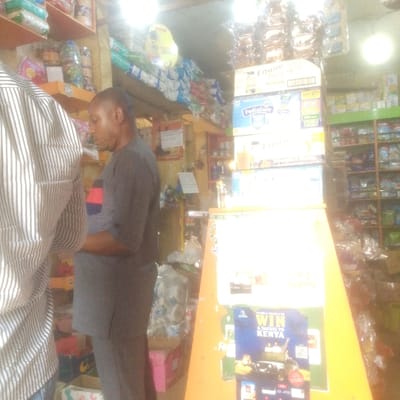 This image depicts the interior of a provision shop or small supermarket located in Abakaliki, Nigeria. The scene is illuminated by bright overhead artificial lighting, with some glare visible.
Two male individuals are present. In the immediate foreground, on the left, the upper back and right arm of a person wearing a vertically striped shirt (white and dark stripes) are partially visible. To their right, positioned centrally, a dark-complexioned male with short, dark hair stands in profile, facing left. He is wearing a dark grey short-sleeved tunic featuring a horizontal red and blue stripe on the left chest, paired with matching dark grey trousers. His head is tilted downwards, suggesting he is looking at something on the floor or a lower display.
The shop environment includes multiple wooden shelves lining the walls, densely stocked with various packaged consumer goods, including snacks, toiletries, and food items, many contained in clear plastic bags or wrappers. A prominent orange display stand stands in the mid-ground, featuring a vertical stack of cardboard boxes labeled "Ensure Plus" and "PediaSure." Below these boxes, a promotional poster for a "WIN A SAFARI TO KENYA" competition is attached to the stand. The ceiling appears to be constructed from simple wooden planks or panels. No direct interaction between the two visible individuals is observed.