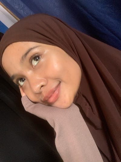 A close-up, waist-up shot captures a young woman wearing a brown hijab. Her right hand, adorned with a light purple sleeve, rests on her chin, supporting her head as she looks upwards and to the left. Her skin appears dewy and reflects light, suggesting a subtle makeup application. The background is a dark, draped fabric, possibly a curtain, with a hint of blue visible on the top right. The scene is indoors, with soft lighting. The location is not specified in the image itself.