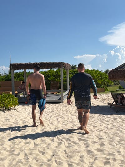 Two men walk across a sandy beach under a clear blue sky with scattered clouds. The man on the left is shirtless, wearing patterned swim trunks, and carrying a blue towel. The man on the right wears a long-sleeved black rash guard and patterned swim trunks. In the background, a thatched-roof structure provides shade, and dense green foliage lines the area behind it. Further to the right, a lounge chair with a person reclining on it is visible. The scene appears to be a resort or beach area. The location is not specified in the provided image.