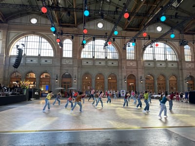 The image depicts a performance by a dance group on a wooden stage in a large indoor venue with arched windows and ornate architecture. The venue appears to be Zurich Main Station (Hauptbahnhof Zürich), a historic building in Zurich, Switzerland, known for its grand hall. Several dancers, dressed in casual attire, are actively engaged in synchronized movements across the stage. In the background, an audience is visible, some seated at tables and others standing. The environment includes stage lighting, speakers, and decorative elements such as hanging colored lights, contributing to the festive atmosphere of a public event.
