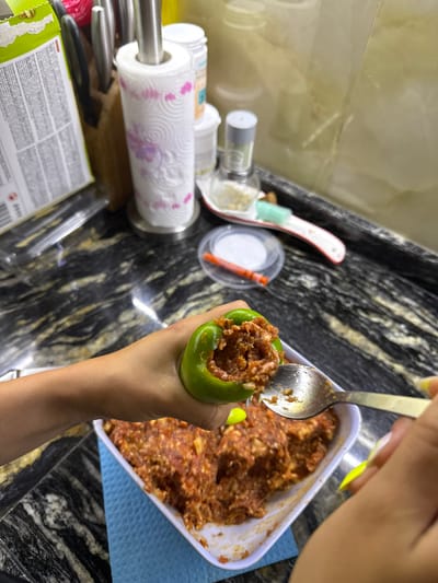 The image shows a person's hands stuffing a green bell pepper with a meat mixture. The bell pepper is being held over a white dish containing more of the meat mixture. A spoon is being used to fill the pepper. The background shows a kitchen counter with various items, including a paper towel holder, jars, and utensils. There are no visible indications of the location being Zornitsa, Bulgaria.