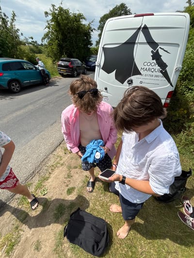 A group of three people are seen outside, beside a road with parked cars. One person wears a pink shirt and dark shorts and holds a bottle to their bare chest with a blue cloth. Another wears a white shirt and dark shorts, holding a phone. The third person is partially visible in the foreground, wearing shorts. A van with "COOPERS MARQUEES" is parked next to them, and various bags and shoes are scattered on the grass.