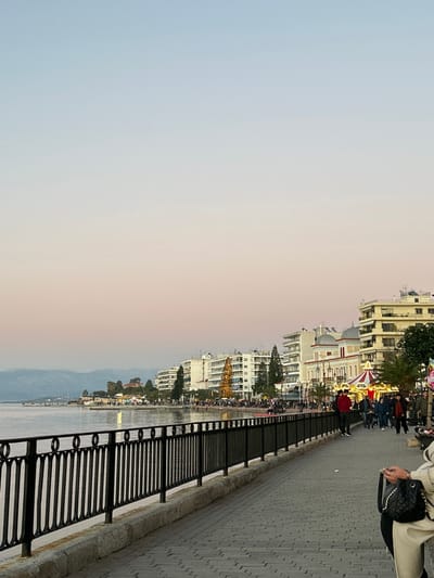 This image captures a vibrant waterfront promenade in Chalkida, Greece, bathed in the soft light of what appears to be either dawn or dusk. The sky transitions smoothly from a pale blue on the left to a warm, soft pink and orange hue towards the right.
On the left, a calm body of water, likely the Euripus Strait, stretches out, reflecting the gentle light. Distant mountains or hills are visible across the water, partially obscured by the soft atmospheric haze.
To the right, a paved promenade, constructed from patterned grey pavers, runs alongside the water. A sturdy black metal railing, featuring a series of circular designs, separates the walkway from the water's edge.
Lining the promenade are several multi-story buildings, predominantly light-colored (beige or white), showcasing various architectural styles, including some with domed roofs. Interspersed among these buildings are lush green trees, including palm trees, adding a touch of natural vibrancy to the urban landscape.
The scene suggests a festive atmosphere. A large, illuminated golden conical structure, likely a decorative Christmas tree, stands prominent near the water's edge. Further along the promenade, a brightly lit carousel with various figures can be seen, hinting at a public event or seasonal celebration.
Numerous people are present on the promenade, enjoying a leisurely stroll. In the immediate foreground on the right, a person wearing a light-colored jacket and carrying a black quilted handbag is partially visible. Further down the promenade, more individuals and small groups are walking or gathering, contributing to the lively yet relaxed ambiance.
The clear sky, calm water, and soft lighting create a peaceful and inviting setting for a community gathering or evening walk. No visible text is discernible in the image.