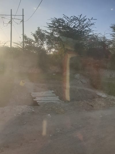 A hazy outdoor scene shows a dirt road bordered by trees and foliage. A set of wooden planks sits along the road's edge. Electrical poles are visible against a pale blue sky, adding a sense of rural infrastructure. The image has a washed-out, ethereal quality, potentially due to sunlight or a lens flare.