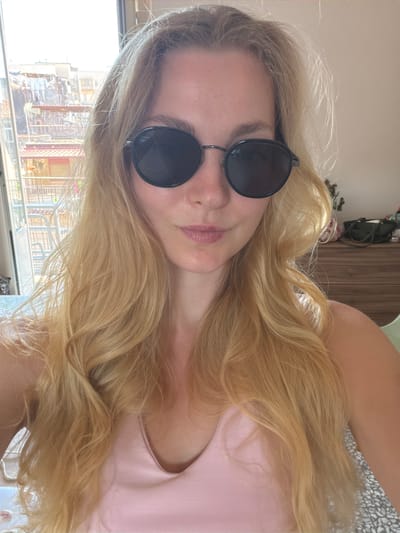 A young woman with long blonde hair is taking a selfie in an apartment. She is wearing sunglasses and a pink tank top. The background shows a window with a view of buildings in Catania, Italy, and a dresser with various items on top. The overall lighting suggests it is daytime.