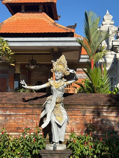 The image captures a Balinese statue adorned with gold detailing, positioned next to a brick wall and verdant greenery with red flowers. The architecture in the background suggests a traditional Balinese temple setting, likely located in Kutuh, Indonesia. Part of the pagoda is seen, along with a palm tree. The statue itself appears to be in a classical dance pose, with one arm extended. A building and roof are visible.