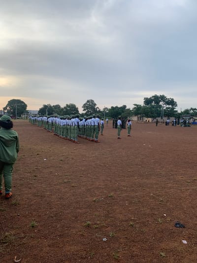 A wide shot depicts a large outdoor gathering on a reddish-brown unpaved field under an overcast sky in Kubi A, Nigeria. Approximately fifty individuals, primarily young adults, are observed standing in a long, single-file formation, facing away from the camera. They are uniformly dressed in light green trousers, white t-shirts, and green caps, consistent with National Youth Service Corps (NYSC) attire. One individual in a similar green uniform, with their back to the camera, stands in the foreground, observing the formation. Further to the right, a smaller group of uniformed individuals stands separately, with one appearing to face the main formation. The ground exhibits sparse patches of green grass and scattered small debris, including a black plastic bag and white scraps. The background features a line of green trees, beyond which indistinct buildings and temporary structures are visible.