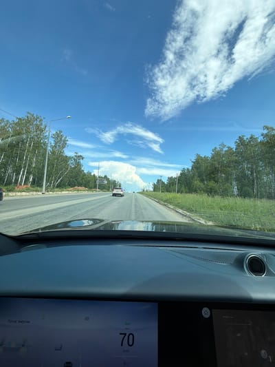 The image is a first-person view from inside a vehicle, looking out at a road scene on a sunny day. The sky is a bright blue with scattered white clouds. A paved road stretches ahead, with trees and green grass lining its sides. In the distance, several cars are visible on the road. The vehicle's dashboard is in the foreground, displaying a digital speedometer reading 70 km/h and other information. The overall environment appears to be a suburban or rural area, possibly on the outskirts of Zapadny, Russia, given the context. The road is well-maintained, and the vegetation suggests a temperate climate. There are streetlights and power lines visible along the roadside. The scene depicts typical daytime driving conditions.