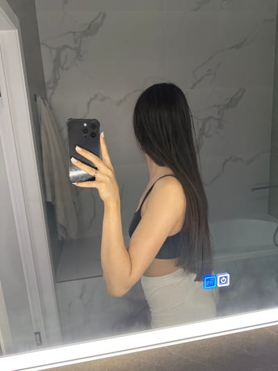 A person is taking a mirror selfie in a bathroom. The person is standing with their back to the camera, wearing a black tank top and a light-colored skirt. They are holding a black smartphone in their right hand, taking the photo. The mirror reflects the person and the bathroom environment, which includes a marble-tiled wall, a towel hanging on a rack, a bathtub, and a smart mirror with illuminated icons. The image does not provide any information about the location in the city of Estachi, Russia.