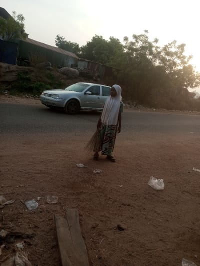 A woman in a headscarf stands on a dirt shoulder next to a paved road, holding a broom. A silver car is parked on the road behind her. Litter is scattered on the ground near the woman. In the background, there are trees and some buildings on a hillside, obscured by haze. The lighting suggests it is early morning or late afternoon.