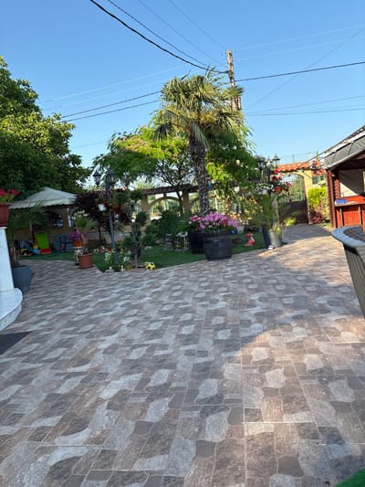 The image shows a residential courtyard in Slanchevo, Bulgaria, under a clear blue sky. The courtyard is paved with large, irregularly shaped tiles in shades of brown, grey, and beige, arranged in a mosaic pattern. In the foreground, a portion of a dark grey woven chair is visible on the right.
The midground features a lush garden with various plants and decorative elements. A tall palm tree stands prominently, surrounded by smaller trees and shrubs. Several potted plants, including those with bright pink flowers, are placed in dark planters, one of which is barrel-shaped. A traditional lamppost with multiple lights stands near the center of the garden. Behind the lamppost, a wooden fence encloses a section of the yard. Further back, an arched gateway with red-tiled roofing leads to another area, possibly a street or driveway. A small, colorful children's slide is visible on the left side of the image, near a white gazebo. A yellow soccer ball rests on the grass in front of a small garden gnome.
The background reveals more of the residential structures, including a red-sided building with a covered patio and a corrugated metal roof. Overhead, power lines crisscross the sky. The overall scene depicts a well-maintained outdoor living space, likely a backyard or patio area of a home in Slanchevo.