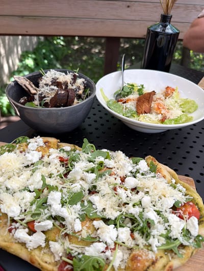 The image shows a table setting with three dishes. A white bowl contains a Caesar salad with croutons and shrimp. A gray bowl contains a salad with dark croutons, possibly mushroom-based. A flatbread pizza with arugula, tomatoes, mozzarella, and shredded cheese sits on a wooden board. A black bottle is also visible. The location may be in or around Ragaciems, Latvia.