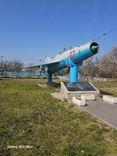 This image depicts a static display of a Mikoyan-Gurevich MiG-21 jet fighter aircraft in Dobroslavtsi, Bulgaria. The aircraft is painted light gray on top and light blue on the underside, with a prominent red numeral "07" on its fuselage and a green emblem on its vertical stabilizer. It is mounted on two light blue cylindrical pedestals, angled slightly upward with its nose cone, which is green, pointed towards the upper right. A long, slender pitot tube extends from the nose. Two missile-like objects are attached under its wings.
The aircraft is situated within a grassy park area. In front of the aircraft, a dark gray paved path runs horizontally. Adjacent to this path, directly below the aircraft's forward section, is a stone-clad base supporting a dark, rectangular plaque. Three rectangular white planters are positioned on the paved path near the plaque.
In the background to the left, a chain-link fence with a blue lower section is visible, along with multiple tall light poles. Beyond the fence, bare deciduous trees and a large, light-colored structure can be seen. To the right background, a dense cluster of bare deciduous trees stands, and a colorful arrangement of vertical multi-colored slats is partially visible. The sky is clear and bright blue, indicating sunny weather. No people are present, and no actions or interactions are depicted.