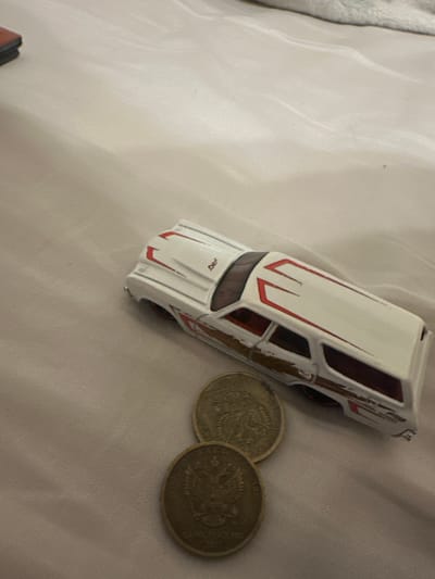A white toy station wagon with red racing stripes and brown accents rests on a textured beige surface, possibly a blanket. Two round, metallic coins, each bearing the image of a double-headed eagle, lie to the lower left of the toy car. The coins appear to be Russian currency. The overall scene is dimly lit and out of focus, suggesting an indoor setting, possibly a home. No people are present. The location is identified as Malinovka, Russia.