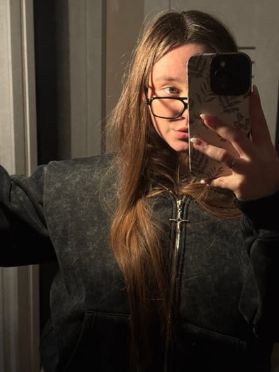 An indoor mirror selfie taken in the city of Prikubanskiy, Russia, depicts a female individual from the chest up. She has long, straight, reddish-brown hair parted in the middle, cascading over her shoulders and chest. The subject wears black-rimmed glasses, with her left eye, part of her nose, and upper lip visible. The right side of her face is obscured by a smartphone held by her right hand. The phone features a light-colored case adorned with dark botanical patterns. Her right hand displays a thin ring on the ring finger and nails manicured with light tips. She is dressed in a dark gray, acid-washed-style hooded sweatshirt with a prominent metallic zipper, the pull of which is shaped like an inverted cross or an ankh. Strong, directional lighting illuminates the subject from the right, creating highlights on her hair and face while casting shadows. The background appears to be an interior space, with a vertical light-colored panel or door frame visible on the left side of the frame.