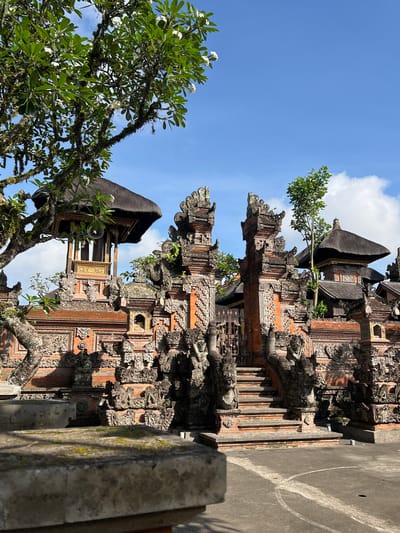 The image shows a detailed view of a Balinese temple structure in Ubud, Indonesia. The structure is heavily adorned with intricate carvings. A tree with white flowers frames the left side of the shot. The sky is blue with scattered clouds in the background. The scene is viewed from a low angle, highlighting the height of the temple.