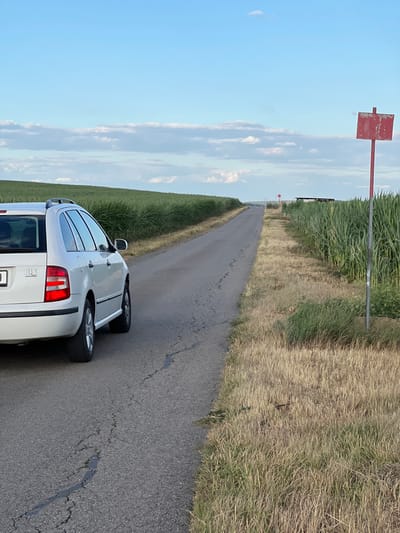 A white station wagon is parked on the right side of a paved road in Měnín, Czechia. The car is facing away from the camera, with its rear to the left. The road is narrow and appears to be in a rural area, bordered by a field of tall green crops on the right and a similar field on the left. The sky is blue with scattered white clouds. A red rectangular sign on a pole is visible in the field to the right of the road. The overall scene depicts a quiet, possibly agricultural, landscape under a partly cloudy sky.