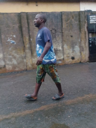 A single adult male is depicted walking from left to right on a damp, dark paved surface. The subject is of dark complexion, with short, dark hair, and is oriented in profile, looking forward. He wears a dark blue short-sleeved t-shirt featuring a light blue and white graphic on the lower front. His lower body is clad in knee-length shorts with a vibrant, multi-colored pattern in shades of green, orange, black, and yellow. He wears dark slide-style sandals on his feet. His posture indicates motion, with his right leg extended forward and left arm slightly forward.
The background consists of a textured, weathered wall, primarily grey and beige, extending horizontally behind the individual. Above this wall, a narrower band of faded orange-yellow structure is visible. To the far right, a dark metal gate with faint, illegible markings is partially visible. The paved ground shows streaks of water and subtle reflections. The scene is an outdoor urban or semi-urban environment in the city of Agani, Nigeria, captured under diffuse lighting conditions. The image exhibits slight motion blur, particularly on the moving subject and the ground.