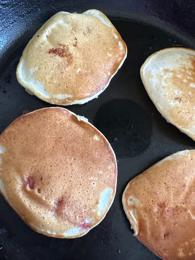 Four golden-brown pancakes are cooking in a dark skillet. The pancakes are circular and have a slightly uneven texture with small bubbles on the surface, indicating they are in the process of being cooked. Some of the pancakes have reddish specks, possibly from fruit mixed into the batter. The skillet appears to be black and has a slightly oily surface. The lighting suggests an indoor setting, possibly a kitchen, and the image is a close-up shot, focusing solely on the pancakes and the skillet. There is no visible text or any contextual information about the time of day, weather, or emotions. The scene depicts the activity of cooking pancakes.