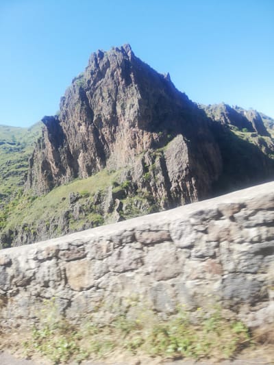 This is a photograph of a mountainous landscape in Tmogvi, Georgia. In the foreground, a blurred stone wall runs horizontally across the image. Beyond the wall, a steep, rocky mountain face dominates the center of the frame, with patches of green vegetation clinging to its slopes. The mountain is rugged and appears to be composed of layered rock formations. The sky above is a clear, bright blue. The overall impression is of a remote and natural environment.