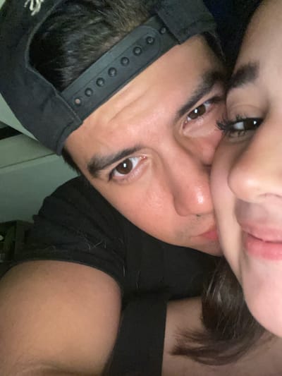 The image displays a close-up of two individuals in an intimate pose within an unidentifiable indoor setting. The scene is dimly lit.
The primary subject, a male, occupies the left and central portion of the frame. He is wearing a black baseball cap backward, revealing the black plastic snapback adjuster. His hair is dark and short. He has dark brown eyes, a prominent nose, and appears to be looking directly at the camera. He is wearing a black short-sleeved shirt, on which several light-colored fibers or lint are visible near his right shoulder and upper arm. His right arm and shoulder are prominent in the lower left.
The second subject, a female, is positioned to the right of the male, with only the right side of her face visible. Her right eye has long, dark eyelashes, and she possesses a slight smile. Her dark brown hair cascades along her right cheek and neck. The male's face is pressed against her right cheek/nose area, indicating close physical proximity. Both individuals exhibit features consistent with Hispanic ethnicity.
The background is dark and out of focus, providing no discernible details about the specific room or surroundings. A faint, lighter-toned object or surface is vaguely visible in the upper left background.
The image provides no visual information to determine its location within the city of Coatzacoalcos, Mexico.