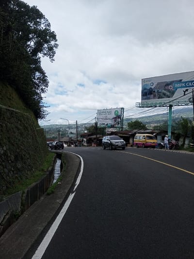 The image depicts a daytime scene on a curved asphalt road in the city of Phoxay, Laos, under an overcast sky with patches of white clouds. To the left, a steep, vegetated hillside with dark green trees is supported by a stone retaining wall. A concrete drainage ditch containing water runs alongside the road at the base of the wall. The asphalt road curves to the right, marked by a solid white line on its left edge and a dashed yellow line on its right.

A dark grey SUV is in motion, traveling from the right side of the frame towards the left. Further along the curve, a queue of dark-colored vehicles is visible in the distance. On the right side of the road, a yellow and pink mini-van is parked on the shoulder, next to a red motorcycle. Two individuals are standing near these parked vehicles; one is wearing a light-colored long-sleeved shirt and pants.

Numerous overhead power lines and utility poles are present. Two large billboards are prominently displayed: a lower one on the left advertises "LEMBAH HIJAU RESORT HOTEL CILOTO" with an accompanying building image, and a higher one on the right advertises "BBPK CILOTO" with architectural designs. Beyond the roadside structures, a valley extends with distant green hills and scattered buildings.