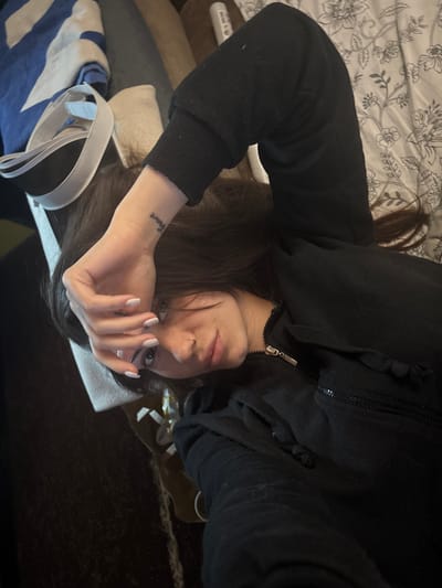 An indoor portrait captures a person, likely female, lying down, appearing to take a selfie in Belopoptsi, Bulgaria. The image is rotated 90 degrees clockwise, showing the person's head to the left. They have long, dark hair and are wearing a black hooded jacket with a zipper and drawstrings. Their left hand, featuring light-colored nail polish with small dots, is raised and partially obscuring the face, with fingers slightly spread. A small tattoo reading "brave" is visible on the inner wrist of this hand. The person has a nose piercing and looks directly at the camera with a neutral gaze. Behind them, a white and grey floral patterned bedding or blanket is visible, suggesting the person is on a bed. Other objects in the background include blue and white striped fabric, a white strap, and indistinct dark surfaces. The lighting is soft and consistent with an indoor setting.