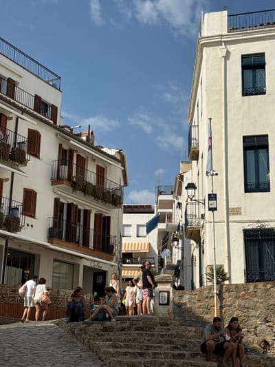 The image captures a daytime scene on Carrer de Bofill i Corona in Calella de Palafrugell, Spain, under a clear blue sky with scattered white clouds. The scene is dominated by two light-colored, multi-story buildings flanking a pedestrian street that ascends towards the background.
The building on the left features white facades, multiple levels of balconies with black metal railings, and dark brown louvered shutters. Some balconies display hanging plants. Its ground floor houses a commercial establishment identified by the sign "Calau," with outdoor seating visible. The building on the right, also white, features dark-framed windows and some balconies. A flag pole with a white and blue flag (possibly a Saltire-like design) is attached to its facade, below which a black street lamp and a blue informational sign are present. A street sign clearly labels the location as "CARRER DE BOFILL I CORONA." Further up the street, a white building with yellow and white striped awnings, partially obscured, is visible, with "SUPERMERCAT" legible on one awning.
The ground surface consists of large, irregular stone steps in the foreground, transitioning to a paved stone surface further up the incline. Approximately 15-20 individuals, primarily adults, are distributed throughout the scene. In the immediate foreground, two individuals are seated on the stone steps to the right. In the mid-ground, a group of three women are seated on the stone steps or an adjacent stone wall on the left. Other individuals are standing and conversing near the "Calau" establishment, while more pedestrians are visible walking or standing further up the street. General activity involves casual pedestrian movement and social interaction.