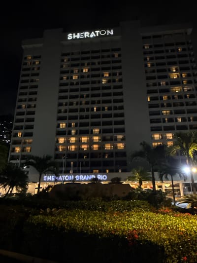 This image presents a low-angle, nighttime exterior view of a prominent hotel building, identified as the Sheraton Grand Rio in Rio de Janeiro, Brazil. The main structure is a tall, multi-story building with a light-colored facade, featuring numerous rectangular windows and balconies across its width. Many of these windows are illuminated with warm yellow light, indicating interior activity.
At the very top of the building, the word "SHERATON" is displayed in large, glowing white letters. Closer to the base of the building, a smaller, similarly illuminated sign reads "SHERATON GRAND RIO."
In the immediate foreground, a dense array of green foliage, possibly a landscaped garden or shrubbery, dominates the lower part of the frame. This vegetation is illuminated from below by artificial lights, highlighting its texture and revealing scattered patches of red flowers. Several tall palm trees are visible, some partially obscuring the lower sections of the hotel facade.
To the far left in the background, a dark hillside or mountain is discernible, dotted with numerous small, faint lights, suggesting distant urban or residential development. The sky above is dark and featureless. No individuals are visible in the scene, and no specific actions or interactions are depicted. The overall ambiance is that of a large, active hotel at night in a tropical, urban environment.
