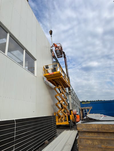 In Stróża, Poland, on a construction site, a scissor lift is positioned against the side of a large, modern building. The building features a light-colored, ribbed exterior wall and several large, rectangular windows. Two workers, wearing safety vests and hard hats, are operating the scissor lift. One worker is in the elevated platform of the lift, appearing to be working on the building's facade. The other worker is at the base of the lift, likely controlling its movement or assisting. Above the scissor lift, a crane hook is visible, suggesting ongoing material lifting operations. In the background, a large blue shipping container with the text "CONTAINEX" is parked, and stacks of building materials, including what appear to be insulated panels, are present on the ground. The sky is partly cloudy.
