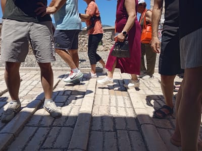 The image shows a group of people walking on a cobblestone street. The focus is on their legs and feet, with some of their torsos visible. They appear to be walking in the same direction. The sunlight casts strong shadows across the ground, suggesting a bright, sunny day, likely midday.
Several individuals are wearing casual clothing suitable for warm weather. One person on the left wears grey shorts and a black t-shirt. Next to them, someone is in blue shorts and a light blue t-shirt. Further to the right, a person in an orange t-shirt and black cropped pants is visible, carrying a black device on their back. Another individual is wearing a long, burgundy dress. One person is wearing dark shorts and a black t-shirt, and another has on a grey and white striped skirt.
Visible footwear includes white sneakers, athletic shoes with red and white accents, black sandals, and simple flat sandals. The cobblestone street is made of irregular, light-colored stones with darker grout lines. In the background, there is a stone wall and some foliage, hinting at an outdoor historical setting. The overall mood seems relaxed and casual, consistent with people on a stroll or sightseeing. No distinct emotions are conveyed due to the cropped nature of the shot. There is some faint text on a poster in the background, but it is illegible. The scene is consistent with a sunny day in a historic European town like Mostar, Bosnia and Herzegovina.