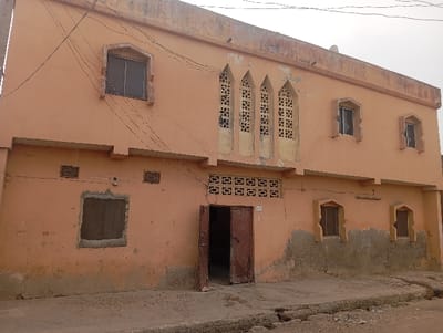 A two-story peach-colored building stands in Gama, Nigeria. The structure exhibits significant weathering, including paint discoloration, peeling, and cracking along the lower first-floor facade. Architectural details include multiple pointed arch windows and central second-floor panels with geometric concrete lattice work.
The ground level features a partially open, double-leaf, brown wooden door. To its left, a rectangular window with a dark mesh screen is positioned beneath two small square vents. To the right of the door, two pointed arch windows with dark mesh screens are observed. A row of square-patterned concrete vents is situated directly above the main entrance.
The second floor includes a pointed arch window with a dark mesh screen on the left. The central section displays three tall, narrow pointed arch panels filled with intricate geometric concrete lattice work. To the right, two additional pointed arch windows show evidence of broken or missing glass panes. Several black electrical wires traverse the facade at the second-story height. The immediate foreground consists of a cracked and uneven concrete pavement. No persons or overt activities are present.